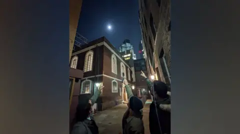 Three men stand outside the synagogue at night and point towards the moon, which appears full. 