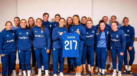 Ipswich Town Football Club The Ipswich Town Women's team are pictured standing together in a group. Natasha Thomas is stood in the middle holding her anniversary kit. The top has her name on the back as well as the number 217 marking her number of appearances. 