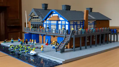 David Johnson Photographic A Lego model of a boathouse showing the ground floor and first floors of the boathouse which is mainly blue and white. Lego figures can be seen on the decks and in front of a river.
