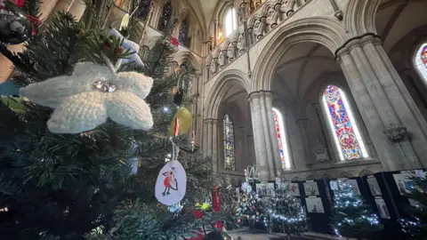 Jo Makel / BBC In the foreground, a white knitted Christmas tree decoration with a bead at its centre, hangs on a fir tree next to a decoration with a drawing of a robin. In the background are four other decorated Christmas trees. The came is angled upwards to show the archways and stained glass windows of beverley Minster