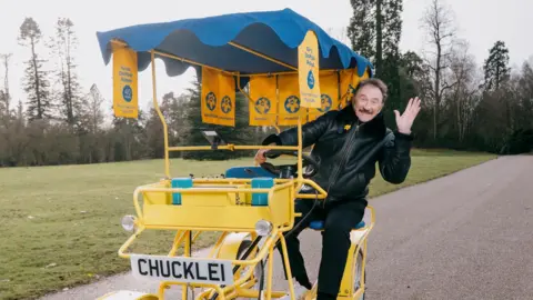 A man in a black jacket and jeans rides a two-seater pedal car in yellow and blue. The number plate on the front reads Chuckle1