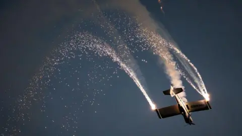 Getty Images A plane flying in the night sky upside-down. It has pyrotechnics coming from the tips of its wings.