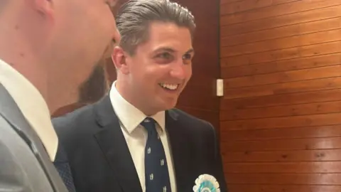 A man smiles wearing a dark blue suit and tie with a light blue rosette on.