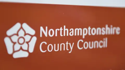 PA Media A former sign for Northamptonshire County Council, which has white writing on a brown background. There is also a white rose on the sign.