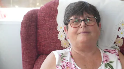 Jamie Niblock/BBC A woman is sitting in a pink armchair. She is wearing a white dress with large pink flowers on it and glasses. She is looking into the camera and smiling. 