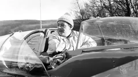 Tom Buist/Mirrorpix/Getty Images Donald Campbell in the Bluebird cockpit. He is wearing a woolly hat and a light zip-up jacket. The craft has a clear canopy, which is opened.