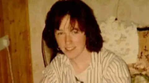 An Garda Síochána A woman, with brown hair, sitting on an old fashioned couch, smiling at the camera. She is wearing a white shirt with blue stripes on it. 