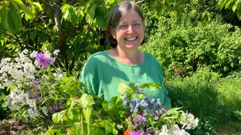 A woman with mid-length greyish hair and wearing a green top stands in the midst of trees and bushes on a sunny day. She is holding long-stemmed flowers, which are white, purple and blue.