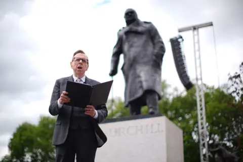PA Media Actor Timothy Spall wears a suit and tie as he reads from an A4 booklet in front of the statue of Churchill in Parliament Square.