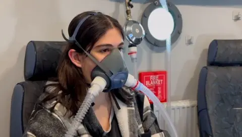 Contributor photo Kaylee is sat looking ahead whilst hooked to an oxygen tank. She has dark hair loose around her shoulder and wears a checked fleece jacket.