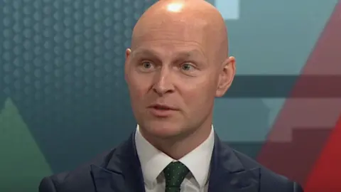 Lib Dem MP Max Wilkinson, a bald man wearing a white shirt and green tie under a dark jacket.