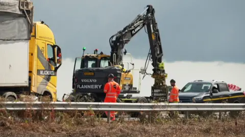 Two men wearing orange high-viz and black helmets are standing next to a metal crash barrier at the side of the road. Next to them is a black pickup truck, a yellow lorry and a black digger. In the foreground you can see brown vegetation. 