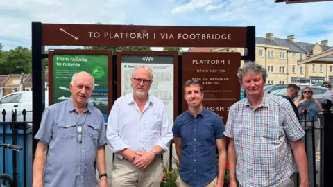 Four men stood by train station platform signs, one which says 'Platform 1 via footbridge'
