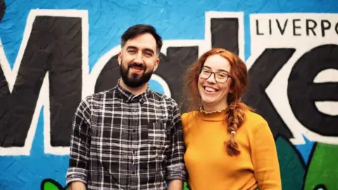 Lee Kent Liam Kelly pictured with Make Co founder Kirsten Little. He is wearing a black-and-white checked shirt and has dark hair and a beard. She has red hair and wears a yellow/orange jumper. Both are smiling.