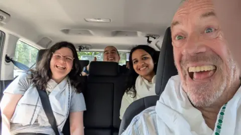 Andy Long taking a selfie inside a car, with Chloe Beer and two course mates in the back. They are all dressed in white and they are all smiling at the camera.