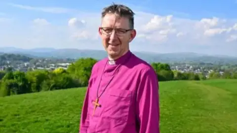 Diocese of Carlisle/Stuart Walker Photography The Right Reverend Rob Saner-Haigh in a bright pink cleric's top and wearing a large crucifix. He is wearing glasses and smiling while standing on a green grassy hill above a town. There are blue skies.