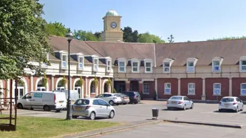 Cars are parked in the car park at the front of the hotel, with a tree to the left of the picture and some hanging baskets to the left hand side of the two storey building, and a sign saying Coach House at the entrance.