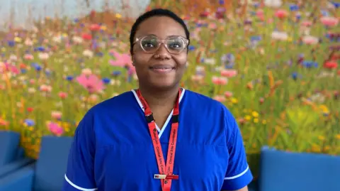 Liz Adeleye wearing glasses and smiling, wearing a blue nurse's outfit. 