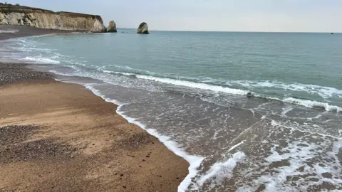 Gigi Photo taken from a sandy beach near to where the water is lapping on to the coast. Cliffs are in the distance, breaking off into columns of rock.