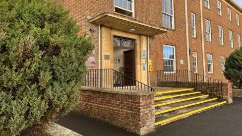 A red brick building with two green bushes in front of it. There are stairs leading up to the building's main entrance. There are a few windows with white frames.