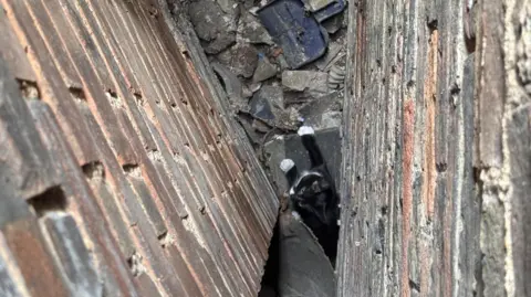 Cambs Fire and Rescue A black and white cat is at the bottom of a gap between brick walls. There is rubble at the bottom of the gap.
