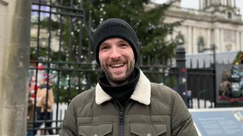 Chris Thompson, a young man wearing a black beanie hat, a black jumper and a khaki colour, fur trim jacket, smiles at the camera. He is standing outside Belfast City Hall with the christmas tree slightly blurred behind him.