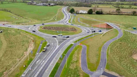 An aerial view of a roundabout with new linking roads around it. One is going over a bridge over the top of one road feeding into the roundabout. Tree saplings have been planted on the banks around the roundabout.