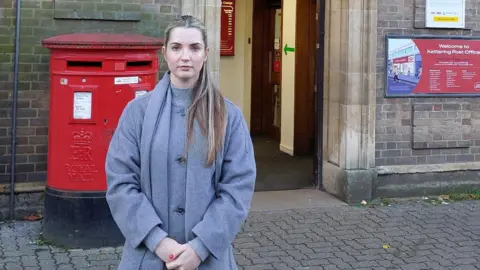 A woman with long blonde hair wearing a grey roll neck jumper, a grey scarf, and matching grey coat stands in front of a post office entrance and two red post boxes. 