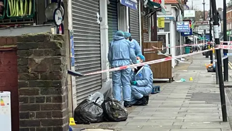 Three people in blue all-in-one suits, gloves, hair covers and masks work within a police cordon outside a shop with shutters pulled down and next to rubbish bags on a high street.