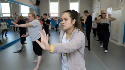 Ian Paine A studio with about 20 people practicing dance moves. There are a mixture of men and women, facing forward, most with a left arm outstretched. one of the walls is mirrored and there are two high windows in the rear wall.