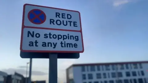 A round white road sign with red edges that reads "RED ROUTE NO STOPPING AT ANY TIME"