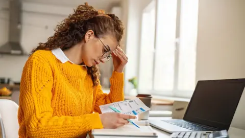 Getty Images Young woman in an orange jumper sits at a desk in a kitchen looking at her bills