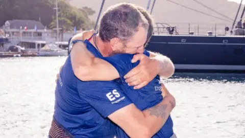 2Stroke.team Karl Austen and Ed Shaw wearing navy t-shirts hugging each other on the jetty at English Harbour in Antigua.