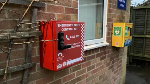 The red bleed kit cabinet and yellow defibrillator box affixed to a brick wall outside the hall. 