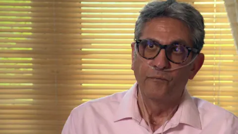 BBC Esse Khanzadeh wearing glasses, a pink shirt, and an oxygen tube around his nose, looks at the camera as he sits down in a room, with drawn blinds behind him 