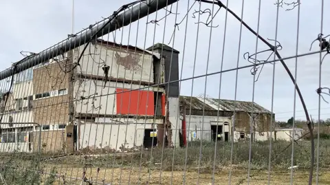 A view of the former Cummins generator factory in Stamford through a wire fence. The ground surrounding the building is overgrown with weeds and plants. Parts of the building have been demolished and several warning signs not to enter the site are visible.