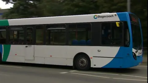 A mostly white single-decker bus, with blue at the front and green at the back, drives on a road. 