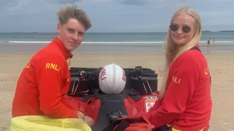 Wilber has short blond hair and is wearing a red lifeguard fleece, Freya has blond long hair and is wearing the same fleece and sunglasses. They are sitting on a lifeguard open top motor car with the beach and sea behind them. They are smiling at the camera