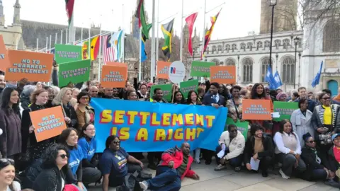 A large group of people including refugees holding banners and flags. They are standing outside Parliament. A large banner reads, Settlement as a Right.