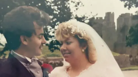 Family Photo Trasi and Mark Coghlan smile at each other, Trasi is wearing a white veil and dress. She has long blond hair. Mark is wearing a black suit jacket with a grey vest and white collar. He has a moustache and brown short hair. Behind the couple a castle is visible between the trees.