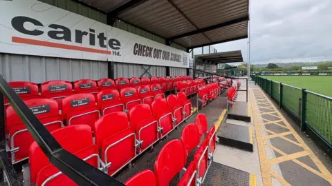 The image shows the football seating area. The seating is covered by a shelter, there are four rows of red chairs. 