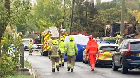 Jenny Kirk/BBC A number of emergency service workers in road in Bedford. They are walking away from the camera to a house that has been destroyed. A number of cars are  in the road, a police tent and a fire engine is in the street. The road has a number of trees and is in a residential area. 