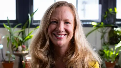 Karen Canfell, wearing a gold top, smiles at the camera, potted plants in the background behind her