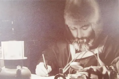 David Bell A sepia photograph of a man wearing a fur hat looking down at a desk. He has icicles in his beard and moustache. He is holding a magnifying glass and looking at papers, while holding a pencil in his hand. A gas lamp in on the desk. The picture gives an impression of him conducting serious work in freezing conditions.
