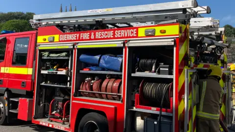 A picture of a Guernsey Fire and Rescue Service truck. It is red and yellow in colour. There is a firefighter wearing uniform and a helmet loading a piece of equipment in the back of it.
