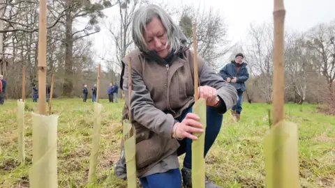 Teresa kneels on one leg as she tends to one of the newly-planted trees, which is at about knee height. She has medium length silver hair and wears a brown coat and gilet, jeans, and boots.