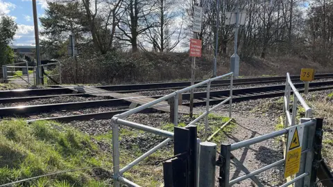 The train line at Pewsey footpath crossing. There are metal gates either side of the train track with caution signs and signage instructing pedestrians to stop look and listen.