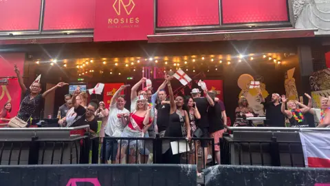 A group of people inside a bar waving red and white England flags. They are smiling with their hands in the air and are singing.