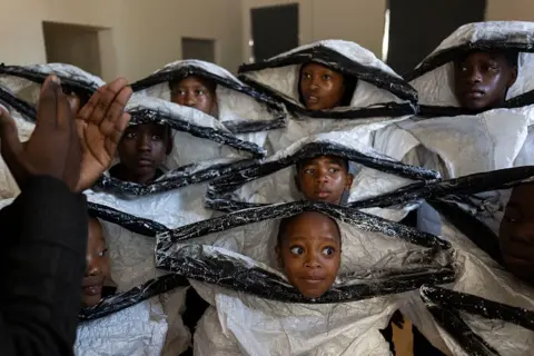 ALET PRETORIUS / GALLO IMAGES / GETTY IMAGES Children in matching white and black outfits huddle together during a rehearsal in a room.