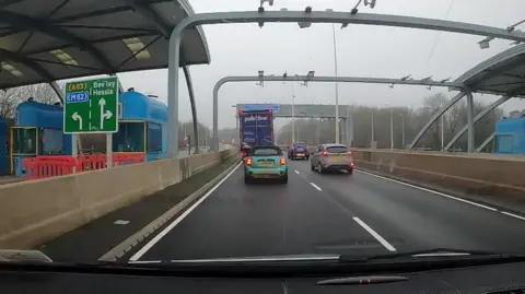 The photo has been taken from inside a car. It's driving on a road, underneath a metal structure which has cameras overhead. To the left are two toll booths, with a green road sign.